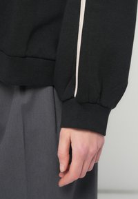 Χέρι που ξεκουράζεται στο πλάι, φορώντας ένα μαύρο μπλουζάκι με μακριά μαν sleeves και άσπρη ρίγα, καθώς και γκρι παντελόνι, κοντινό πλάνο στον μανσέτα του μανικιού και τα δάχτυλα.