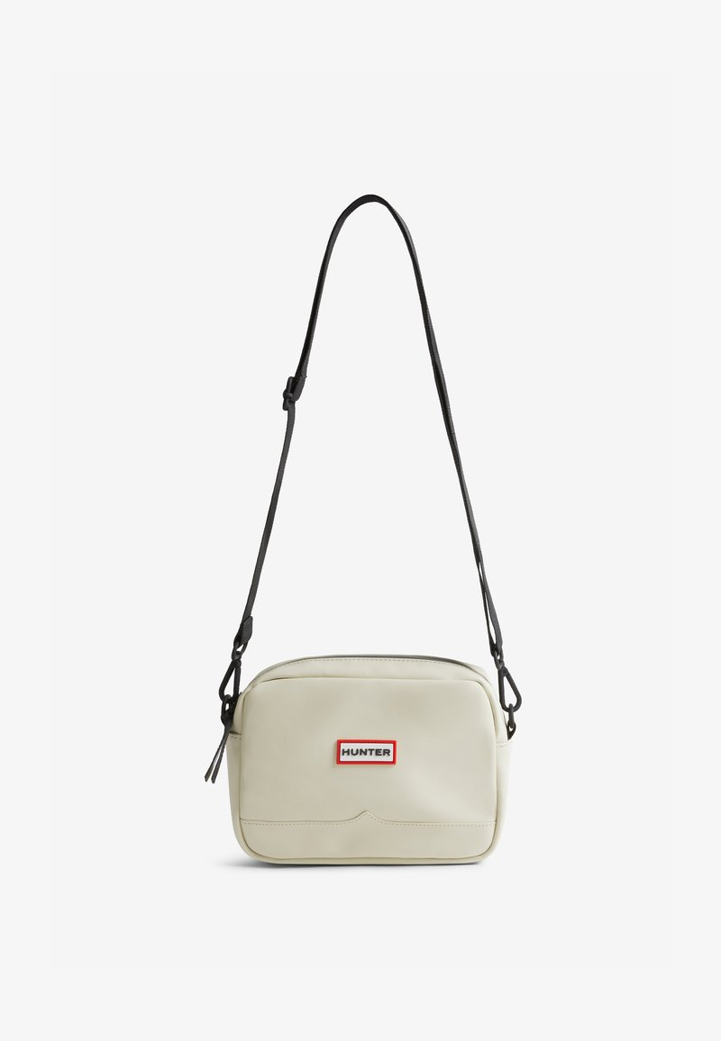 Bolsa de ombro de cor creme, feita de material suave, com fecho de zíper, alça preta ajustável e um emblema com borda vermelha.