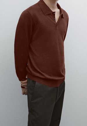 Homme portant un polo en maille marron à manches longues et un pantalon vert foncé, debout avec les mains croisées dans le dos sur un fond uni.