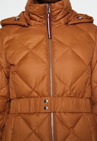 Giubbotto imbottito marrone con trapuntatura, dotato di un colletto alto, zip argentata, vita elasticizzata con bottoni e dettagli in metallo sulle spalle.