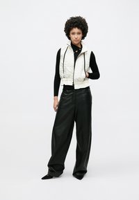 Veste blanche matelassée avec un motif texturé, associée à un haut noir à manches longues et un pantalon en cuir noir ample, complétée par des chaussures noires à bout pointu.