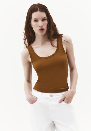 Giovane donna con lunghi capelli castani che indossa un top marrone senza maniche e pantaloni bianchi, con le mani nelle tasche, su uno sfondo bianco semplice.