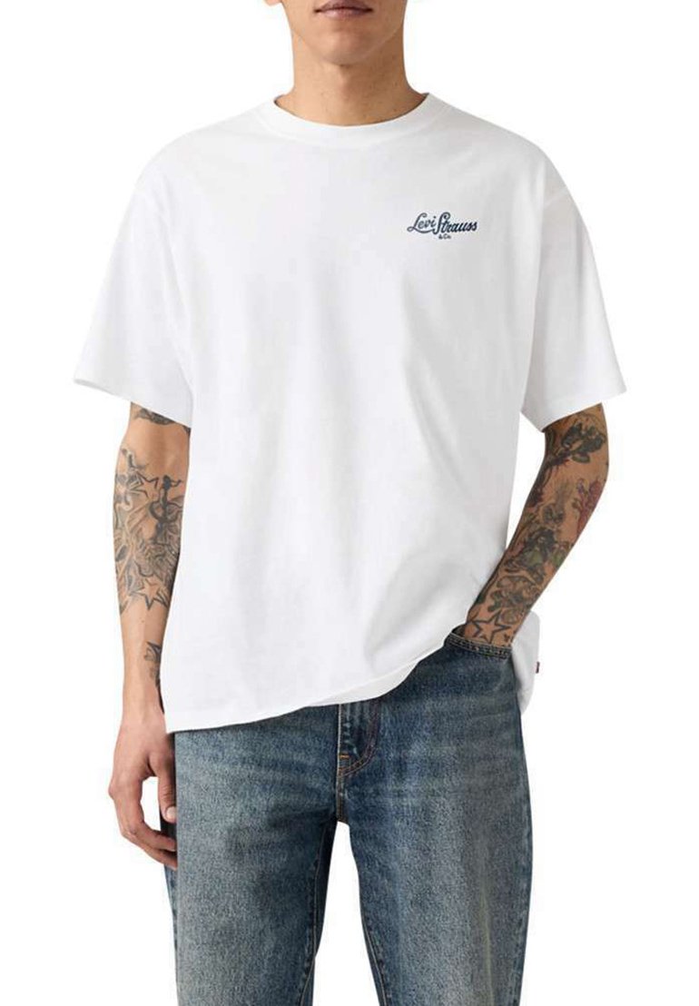 Hombre con camiseta blanca Levi Strauss con logo en el pecho, jeans azules y tatuajes detallados visibles en ambos brazos.