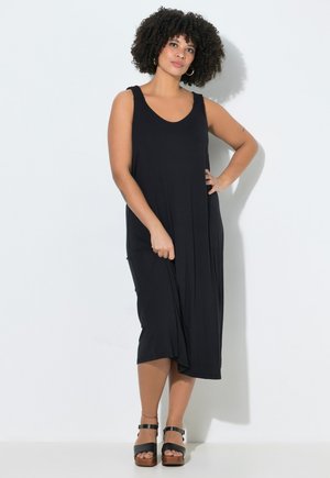 Donna con capelli ricci che indossa un vestito midi nero senza maniche e sandali neri con zeppa, in piedi davanti a uno sfondo bianco.