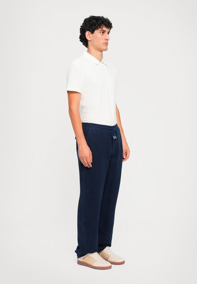 Weißes Polohemd, marineblaue Jogginghose mit Kordelzug und beige Sneakers mit orangefarbenen Akzenten. Glattes Material, lockere Passform und lässiges Design.
