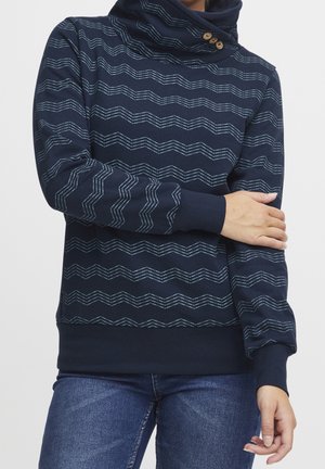 Person trägt einen marineblauen Pullover mit wellenförmigen Streifen und einem hochgeschlossenen, geknöpften Kragen, kombiniert mit blauer Jeans.