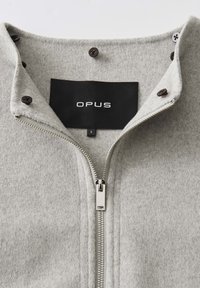 Hellgraue Fleecejacke mit silbernem Reißverschluss und schwarzen Knöpfen, versehen mit einem schwarzen Etikett mit weißem "OPUS"-Text und Größe "S"-Tag.