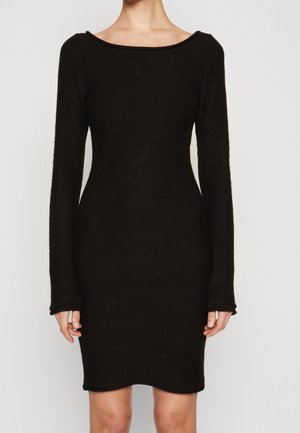 Kobieta ubrana w dopasowaną, czarną sukienkę z długim rękawem, o długości do kolan, z szerokim, zaokrąglonym dekoltem na jednolitym tle.