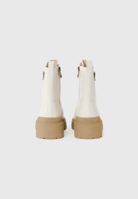 Botas brancas até ao tornozelo com acabamento brilhante, sola robusta em bege e fechos laterais. Apresentam detalhe do logótipo no calcanhar e um design aerodinâmico.