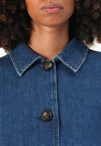 Denimska jakna z ostrim ovratnikom, sprednjim gumbnim zapiranjem in kontrastnim šivanjem. Material je srednje modra, teksturirana denimska tkanina.