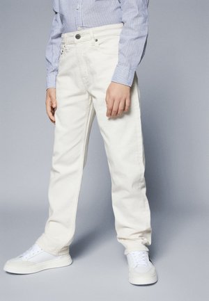 Person iført hvide bukser med lige ben, hvide sneakers og en blå- og hvidstribet langærmet skjorte, stående på en grå baggrund.