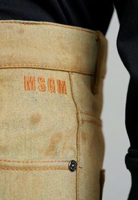 Pantaloni in cotone beige con logo MSGM arancione, caratterizzati da cuciture visibili e un dettaglio in nero sul bottone della tasca. Vista ravvicinata.