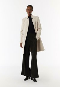 Beige wolmix jas met een rechte snit en drie knopen, gecombineerd met zwarte high-waisted broeken en puntige zwarte hakken.