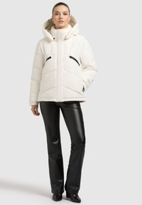Bílá padded bunda s vysokým límcem, černými kapsami na zip a kapucí. Nositelná s černými koženými kalhotami a kotníkovými botami.