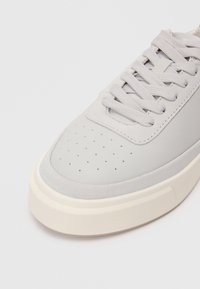 Ανοιχτό γκρι αθλητικό παπούτσι με λείο δέρμα και σουέτ άνω μέρος, στρογγυλή μύτη, διάτρητες λεπτομέρειες στην κάλυψη της μύτης και παχιά κρέμα ρύθμισης από καουτσούκ.