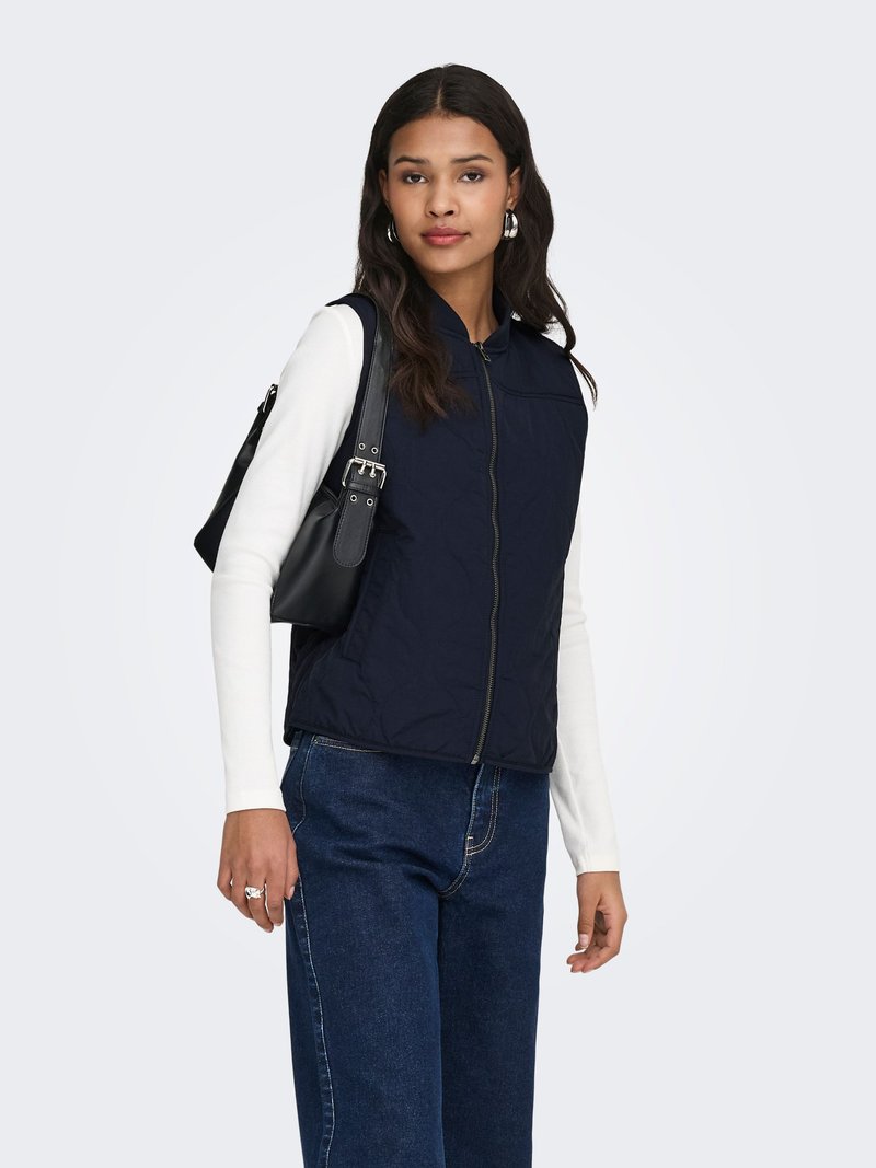 Femme portant un gilet marine zippé sur un haut blanc à manches longues, un jean bleu foncé, des boucles d'oreilles créoles, et portant un sac à bandoulière noir.