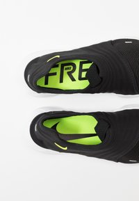 Czarne buty sportowe typu slip-on z siateczkową teksturą, neonowo-zieloną wkładką i białą podeszwą. Posiadają logo Nike i mały żółty akcent.