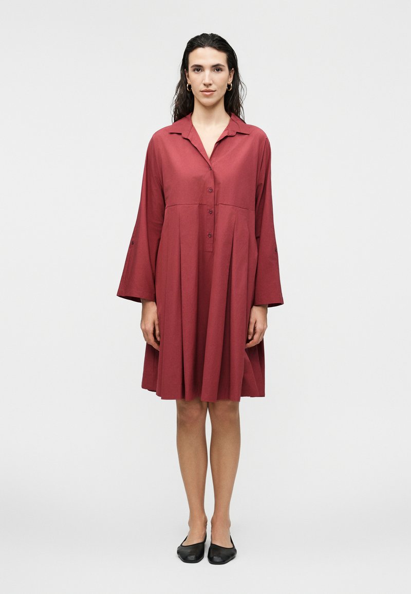 Γυναίκα με μακριά σκούρα μαλλιά που φορά χρυσούς κρίκους σκουλαρίκια και χαλαρό κόκκινο φόρεμα μέχρι το γόνατο με κουμπιά, καθώς και μαύρα φλατ παπούτσια.