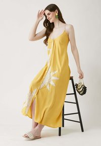 Gelbes Maxikleid mit dünnen Trägern, vorderen Knöpfen und floralem Stickereien. Glatte Textur, seitlicher Schlitz, kombiniert mit leichten Sandalen und einer Perlenhandtasche.