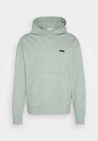 Sudadera de algodón verde menta con un bolsillo frontal, puños acanalados y un logo de etiqueta negra en el pecho. Textura suave y corte relajado.