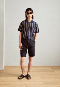 Korte ermer mørkeblå skjorte med hvite og gule striper, svarte shorts og svarte sandaler. Skjorten har krage og knapp-foran design.