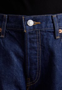 Tmavě modré džíny z denimu s metalickým zapínáním, žlutým prošíváním a pěti poutky na opasek podél pasu.