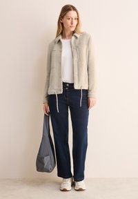 Giacca in pile di colore grigio chiaro, con bottoni, colletto e fondo regolabile con coulisse. Abbinata a jeans blu scuro e sneaker bianche, mentre tiene in mano una borsa grigia.