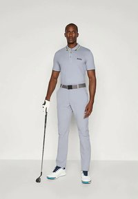 Απαλή γκρι polo μπλούζα γκολφ με γιακά, που διαθέτει κίτρινη και σκούρα μπλε λεπτομέρεια. Συνδυασμένη με απαλές γκρι παντελόνες και λευκά παπούτσια γκολφ. Κρατάει μια ράβδο γκολφ.