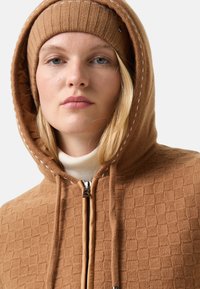 Brun struktureret hoodie med lynlås, der har et mønstret strikdesign og en matchende ribbet beanie. Hvid syning fremhæver hætten.