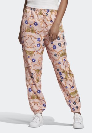 Femme portant un pantalon de jogging rose avec une chaîne dorée et un imprimé léopard, des fleurs bleues, des baskets blanches et un haut blanc, debout avec une main vers le bas.