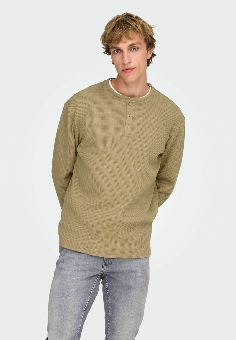 Olivengrøn langærmet henley-skjorte med tekstureret stof, tre-knaps lukning og rund halsudskæring, kombineret med lysegrå jeans.