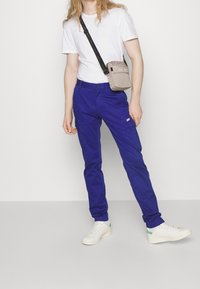 Modré bavlněné cargo kalhoty s bočními kapsami, v kombinaci s bílým tričkem a bílými teniskami se zelenými akcenty, a malou béžovou crossbody taškou.