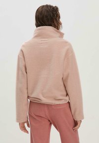 Rosa Fleece-Pullover mit hohem Kragen, lockerem Schnitt und überschnittenen Schultern. Der Stoff hat eine weiche Textur und passt zu einer passenden rosa Hose.