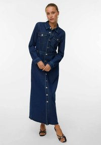 Dolga denimska obleka v temno modri barvi. Ima gumbasto zapiranje spredaj, žepke na prsih in dolge rokave. Teksturni material z oprijetim pasom, rahlo strganje na robu.