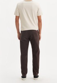 T-shirt en coton beige rayé, manches courtes, associé à un pantalon marron et des baskets noires. Poches arrière visibles sur le pantalon.