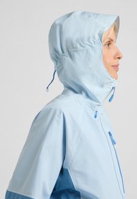 Giacca impermeabile azzurro chiaro con cappuccio, dotata di zip frontale, tasche laterali e un pannello di un blu più scuro su un lato.