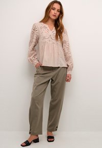 Light pink blouse with eyelet detailing, gathered waist, and long sleeves, paired with loose olive-green trousers featuring button accents.
