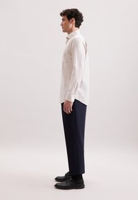 Jasno-beżowa koszula z długim rękawem i kieszonką, noszona z ciemnonawigacyjnymi spodniami i czarnymi butami. Gładka faktura materiału, nowoczesny krój.