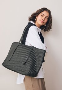 Sac fourre-tout noir en tissu tissé avec accents en cuir lisse, forme rectangulaire et deux anses longues. Caractérisé par un motif texturé et un dessus ouvert.