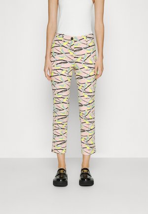 Kvinde iført tætsiddende bukser i ankellængde med lyserød baggrund og mønster af gule fugle på grene, kombineret med sorte loafers og hvid top.