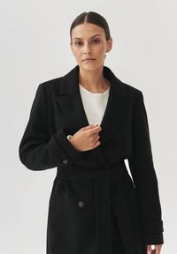 Černý kabát z vlněné směsi s klopovým límcem, dvouřadým zapínáním a páskem v pase. Disponuje velkými knoflíky a předními kapsami. Hladká textura.