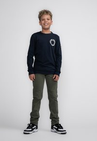 Camicia a maniche lunghe blu marina con grafica bianca, pantaloni verde oliva, sneakers in bianco e nero, in piedi su uno sfondo bianco.