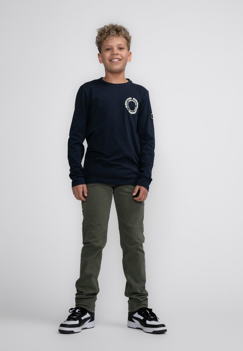 Camicia a maniche lunghe blu marina con grafica bianca, pantaloni verde oliva, sneakers in bianco e nero, in piedi su uno sfondo bianco.