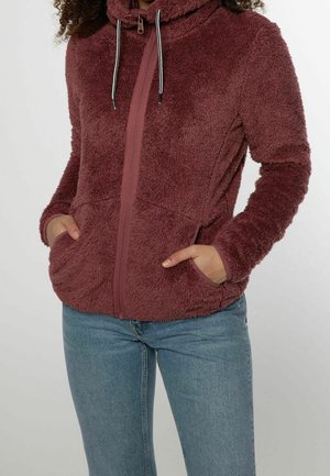 Personne portant une veste bordeaux en tissu pelucheux zippée avec capuche et poches, associée à un jean bleu clair, debout les mains dans les poches.