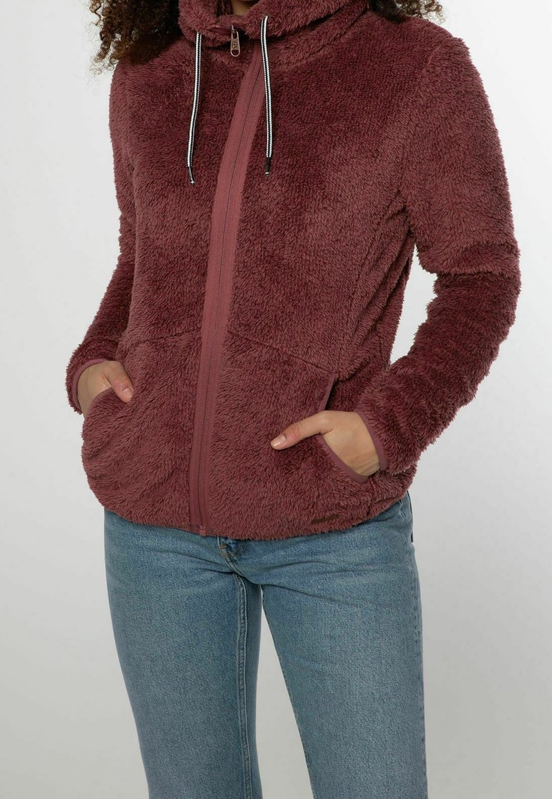 Personne portant une veste bordeaux en tissu pelucheux zippée avec capuche et poches, associée à un jean bleu clair, debout les mains dans les poches.