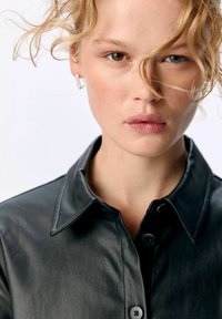 Chemise en cuir noir avec un col pointu, des boutons à l'avant et une texture lisse. Le modèle a des cheveux ondulés et de petites boucles d'oreilles en argent.