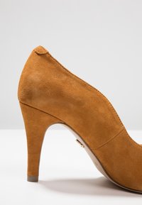 Hnědá semišová vysoká bota na podpatku s špičatou špičkou a štíhlým, zúženým podpatkem. Švy podél stran a malý kovový akcent.