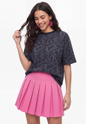 Ung kvinde smiler, iført en mørk mønstret t-shirt, lyserødt plisseret skørt og lyserøde stjerneformede øreringe.