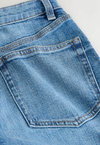 Poche arrière d'un jean en denim bleu avec une couture orange visible et une passants de ceinture sur un fond clair.