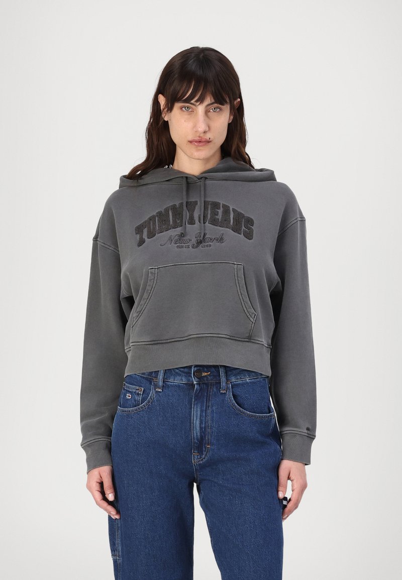 Felpa con cappuccio grigia corta con la scritta "TOMMY JEANS New York" stampata in caratteri scuri e una tasca a marsupio frontale. Indossata con jeans blu.